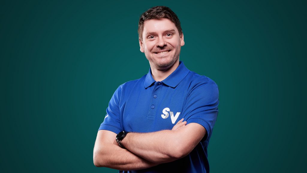Mitarbeiter Alexander freundlich lächelnd in blauem Spandauer Velours Shirt vor einer grünen Wand