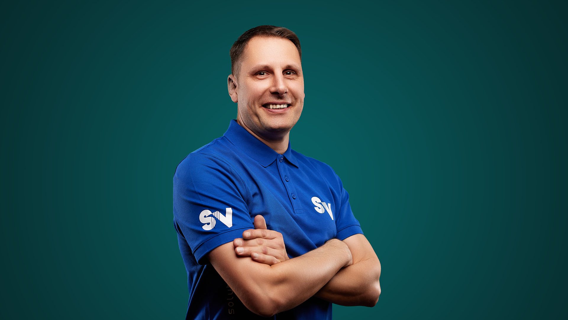 Mitarbeiter Ralf Matthes von Spandauer Velours lächelt mit verschränkten Armen vor einem grünen Hintergrund in die Kamera. Er trägt ein blaues Shirt mit der weiße Aufschrift "SV"