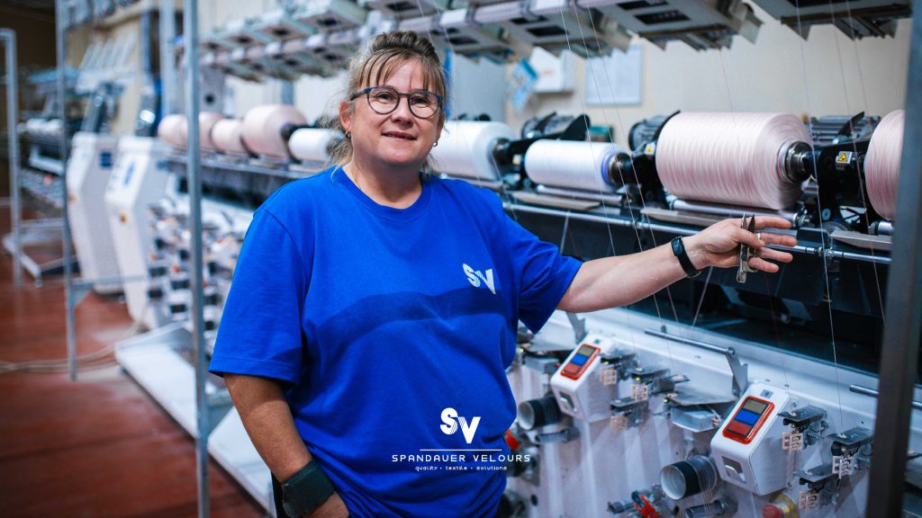 Mitarbeiterin der Firma Spandauer Velours lächelt vor einer Reihe industrieller Garnwickelmaschinen.