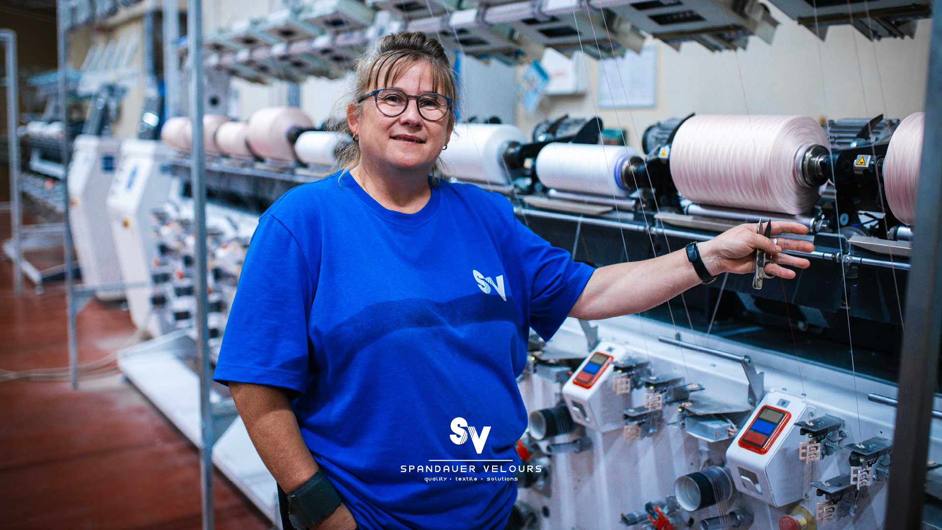 Mitarbeiterin der Firma Spandauer Velours lächelt vor einer Reihe industrieller Garnwickelmaschinen.