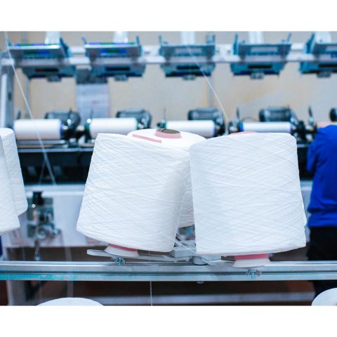 Große weiße Garnspulen im Vordergrund, eine Arbeiterin ist im Hintergrund an der Maschine tätig.