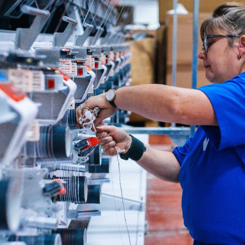 Textilarbeiterin kniet und bindet Fäden an einer Spinnmaschine in einer Fabrik.