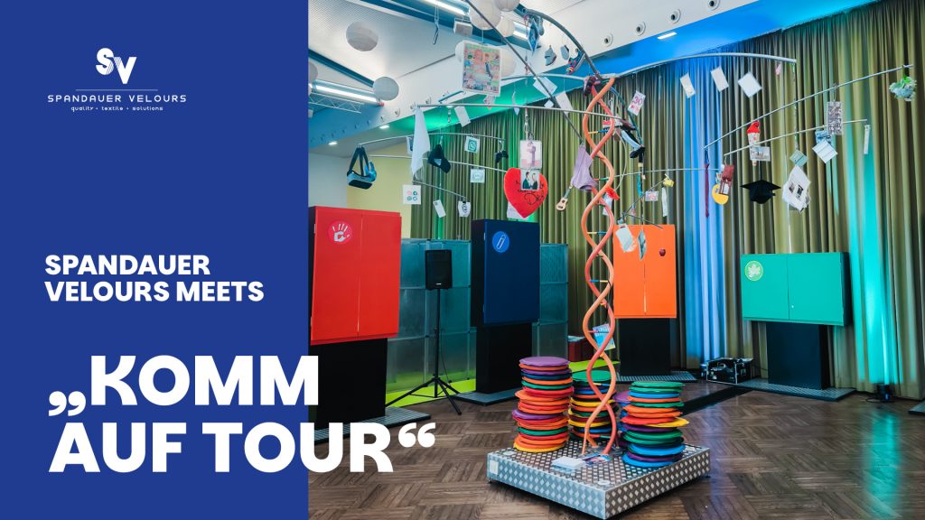 Innenansicht des Standes auf der Berufsorientierungsveranstaltung "Komm auf Tour". Im Zentrum stehen vier bunte, geschlossene Schränke (rot, blau, orange, grün) und eine drahtige Skulptur mit verschiedenen Symbolen, die die Stärken und Interessen der Jugendlichen darstellen. Ein blauer Banner bewirbt das Event: "SPANDAUER VELOURS MEETS 'KOMM AUF TOUR'".