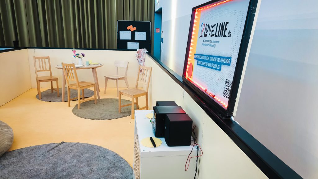 Detailansicht einer ruhigen Ecke bei der Berufsorientierungsveranstaltung. Ein kleiner Tisch mit Holzstühlen, Teppichen und einem Sitzsack bietet eine entspannte Atmosphäre. Rechts beleuchtet ein leuchtender Rahmen das Plakat "LOVELINE.de", ein Informationsangebot zu Liebe, Sexualität und Verhütung, das Teil der umfassenden Jugend-Inhalte der Messe ist.