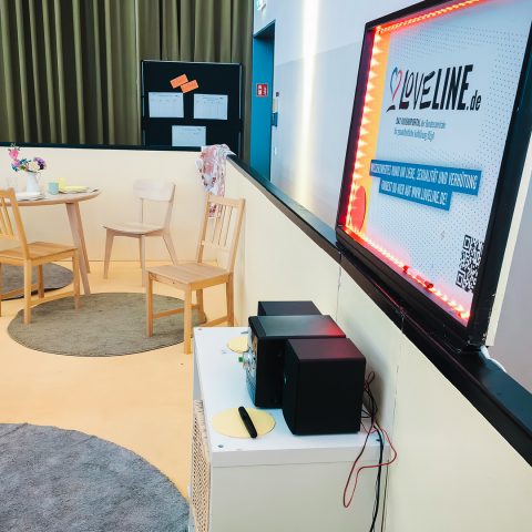 Detailansicht einer ruhigen Ecke bei der Berufsorientierungsveranstaltung. Ein kleiner Tisch mit Holzstühlen, Teppichen und einem Sitzsack bietet eine entspannte Atmosphäre. Rechts beleuchtet ein leuchtender Rahmen das Plakat "LOVELINE.de", ein Informationsangebot zu Liebe, Sexualität und Verhütung, das Teil der umfassenden Jugend-Inhalte der Messe ist.