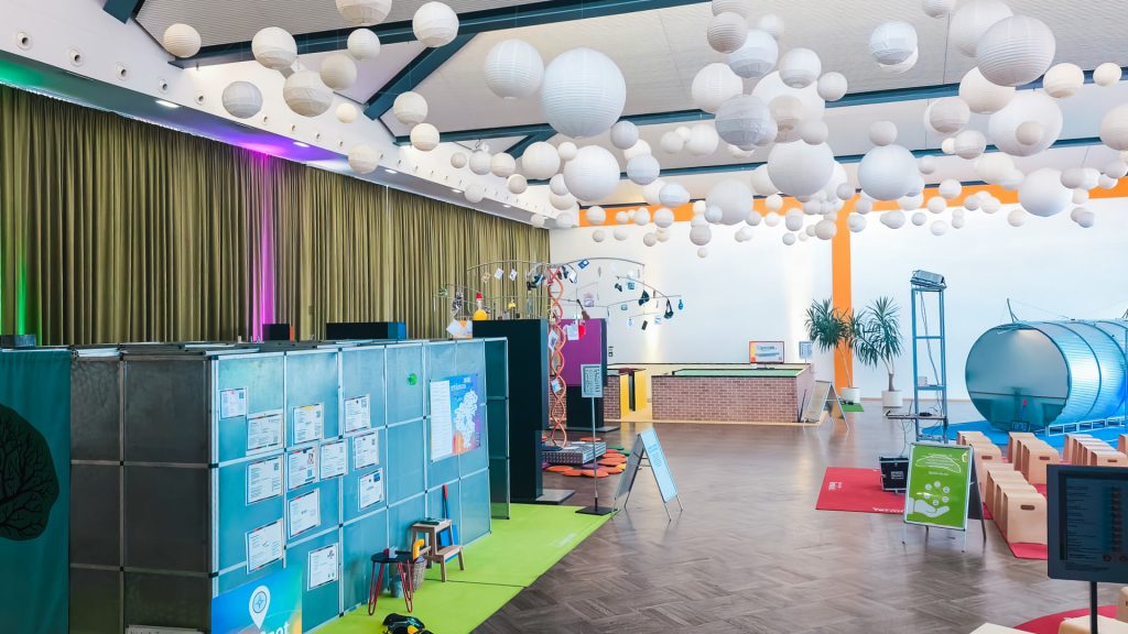 Weitwinkelaufnahme eines großen Veranstaltungsraums bei "Komm auf Tour". Die Decke ist mit vielen weißen Lampions dekoriert. Links ist eine graue, mit Plakaten bedeckte Wand zu sehen, die auf einer grünen Matte steht. Im Hintergrund befindet sich ein großes, silbernes, röhrenförmiges Ausstellungsstück, das Teil der Berufsorientierung für Jugendliche ist.