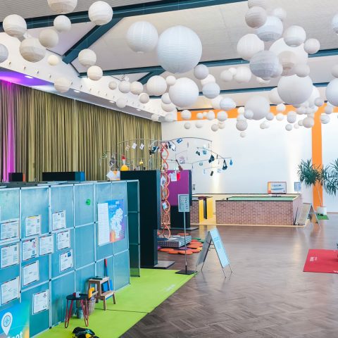 Weitwinkelaufnahme eines großen Veranstaltungsraums bei "Komm auf Tour". Die Decke ist mit vielen weißen Lampions dekoriert. Links ist eine graue, mit Plakaten bedeckte Wand zu sehen, die auf einer grünen Matte steht. Im Hintergrund befindet sich ein großes, silbernes, röhrenförmiges Ausstellungsstück, das Teil der Berufsorientierung für Jugendliche ist.