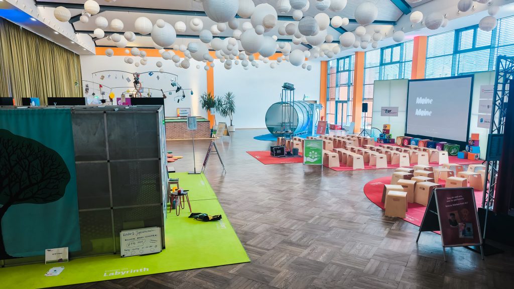 Panoramablick über den Veranstaltungsort der Berufsorientierungsmesse "Komm auf Tour". Die Decke ist übersät mit weißen Lampions. Im Vordergrund sind Sitzbereiche und ein grünes Teppich-Labyrinth zu sehen. Im hinteren Bereich stehen viele kleine Holzstühle vor einer großen Projektionsfläche und einem großen, silbernen Zylinder (Tunnel-Exponat) als interaktiver Teil der Stärken-Findung.