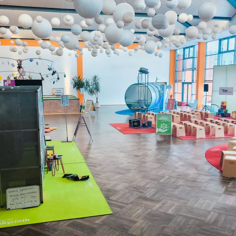 Panoramablick über den Veranstaltungsort der Berufsorientierungsmesse "Komm auf Tour". Die Decke ist übersät mit weißen Lampions. Im Vordergrund sind Sitzbereiche und ein grünes Teppich-Labyrinth zu sehen. Im hinteren Bereich stehen viele kleine Holzstühle vor einer großen Projektionsfläche und einem großen, silbernen Zylinder (Tunnel-Exponat) als interaktiver Teil der Stärken-Findung.