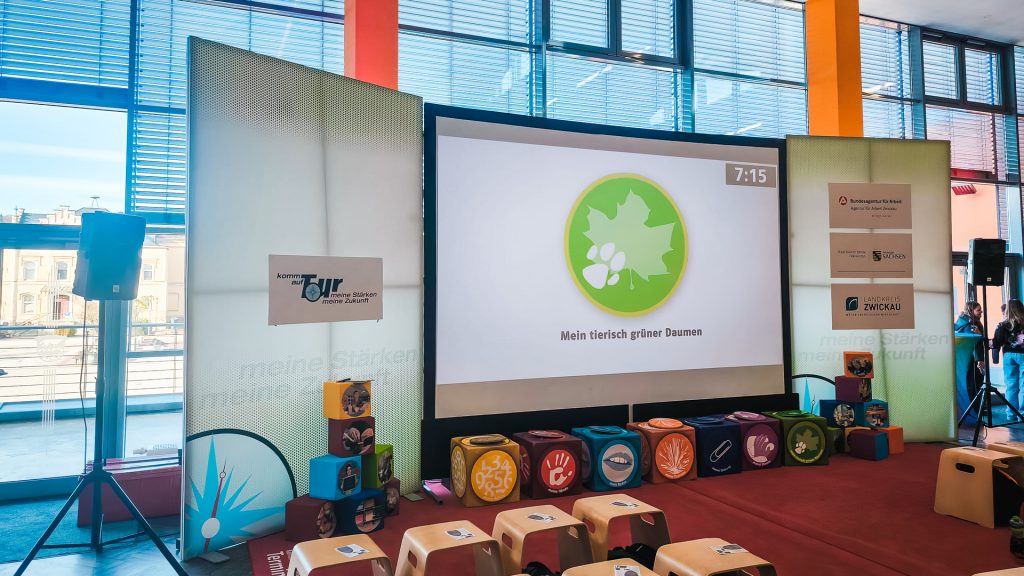 Interaktive Station der Berufsorientierungsmesse "Komm auf Tour" mit einer großen Projektionsfläche, die ein grünes Blatt-Symbol und den Text "Mein tierisch grüner Daumen" (eine der Stärken-Kategorien) zeigt. Vor der Leinwand sind bunte Würfel mit Symbolen für verschiedene Stärken sowie mehrere kleine Sitzhocker für die Teilnehmer aufgestellt.