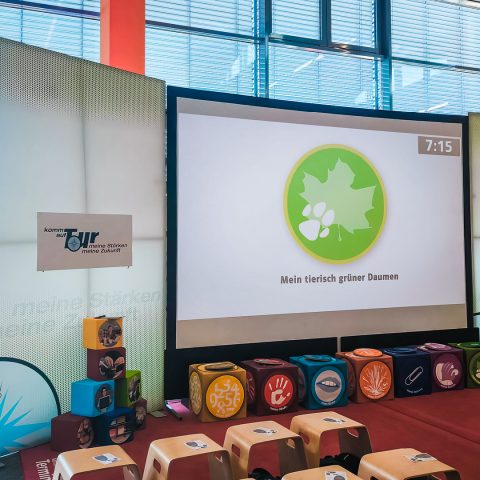 Interaktive Station der Berufsorientierungsmesse "Komm auf Tour" mit einer großen Projektionsfläche, die ein grünes Blatt-Symbol und den Text "Mein tierisch grüner Daumen" (eine der Stärken-Kategorien) zeigt. Vor der Leinwand sind bunte Würfel mit Symbolen für verschiedene Stärken sowie mehrere kleine Sitzhocker für die Teilnehmer aufgestellt.