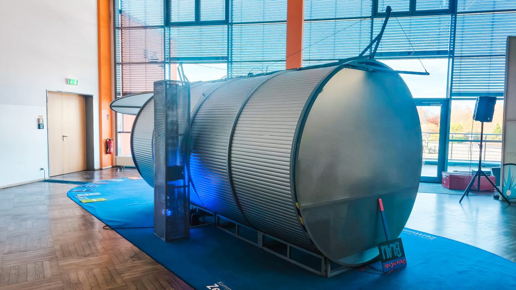 Großaufnahme eines großen, silbernen, zylindrischen Ausstellungsstücks (Tunnel-Exponat) auf der Berufsorientierungsveranstaltung "Komm auf Tour". Das Exponat liegt auf einem blauen Teppich. Es symbolisiert den Weg durch die Berufswahl und die Zukunft der Jugendlichen. Blaue Akzentbeleuchtung sorgt für Stimmung.