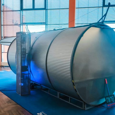 Großaufnahme eines großen, silbernen, zylindrischen Ausstellungsstücks (Tunnel-Exponat) auf der Berufsorientierungsveranstaltung "Komm auf Tour". Das Exponat liegt auf einem blauen Teppich. Es symbolisiert den Weg durch die Berufswahl und die Zukunft der Jugendlichen. Blaue Akzentbeleuchtung sorgt für Stimmung.