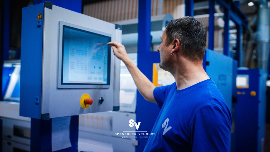 Ein Mitarbeiter mittleren Alters mit einem silbernen Ohrring in einem blauen T-Shirt bedient mit dem Finger ein Touchscreen-Display einer großen blauen Produktions- oder Textilmaschine. Das Display zeigt Steuerungsinformationen. Unten rechts im Bild ist das Logo von "SPANDAUER VELOURS" mit dem Slogan "quality • textile • solutions" eingeblendet.