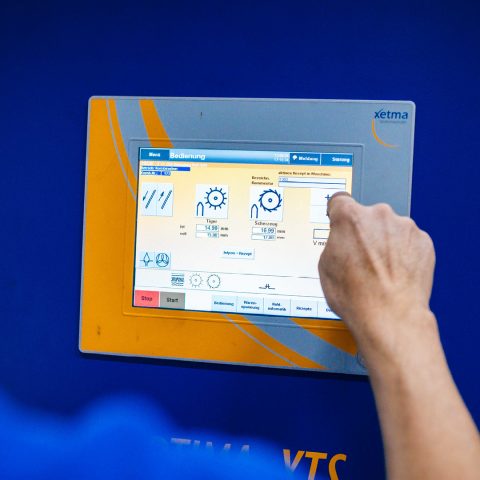 Nahaufnahme einer Hand, die auf ein orange-blaues Bedienfeld einer "xetma VTS" Textilmaschine tippt. Das Display zeigt eine grafische Benutzeroberfläche mit Messwerten und Einstellungen für die Maschinenbedienung, inklusive der Menüpunkte "Bedienung", "Meldung" und "Störung".