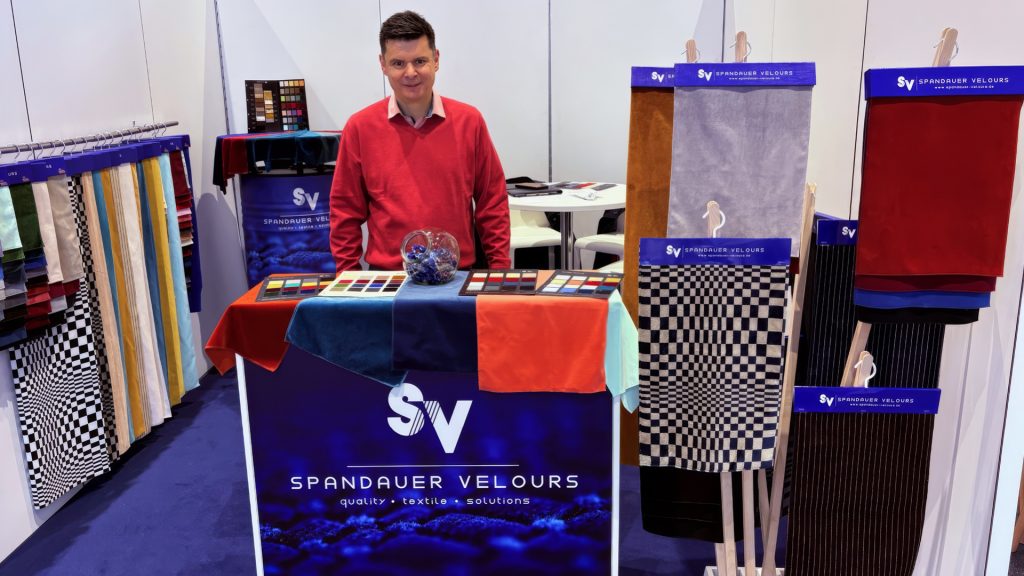 Ein Mann in einem roten Pullover steht lächelnd an einem Messestand von Spandauer Velours, der hochwertige Velours-Stoffe präsentiert. Auf dem Tresen vor ihm liegen Farbmuster und eine Schale mit Deko-Steinen. Rechts und links sind verschiedene Stoffmuster – darunter ein schwarz-weißes Schachbrettmuster, ein braun-schwarzes Streifenmuster und ein leuchtend roter Velours – auf Ständern ausgestellt. Der Stand ist in dunkelblau gehalten und trägt das Logo SV Spandauer Velours | quality • textile • solutions.