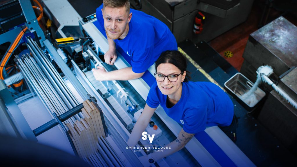 Zwei Mitarbeitende der Spandauer Velours in blauer Arbeitskleidung blicken lächelnd von einer Textilmaschine in der Weberei nach oben in die Kamera.