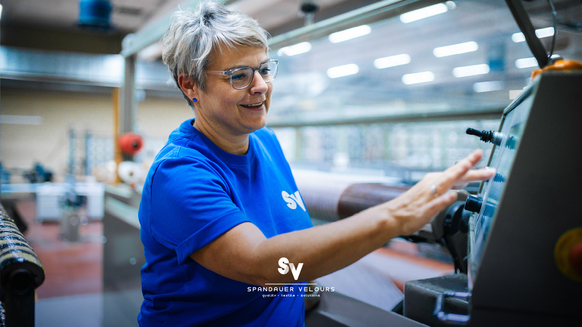 Erfahrene Mitarbeiterin der Spinnerei Spandauer Velours bedient ein digitales Steuerungspanel einer Textilmaschine in der Produktionshalle.