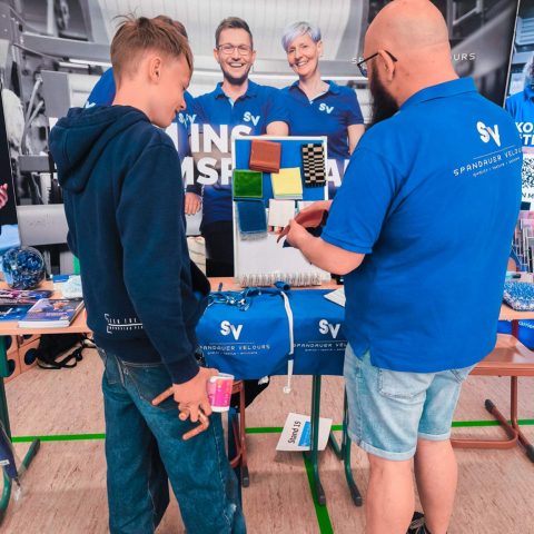 Ein Mitarbeiter von Spandauer Velours im blauen Firmen-Poloshirt zeigt einem Schüler verschiedene Stoffproben und Textilmaterialien an einem Displayboard am Messestand (Stand 19) in Lugau.