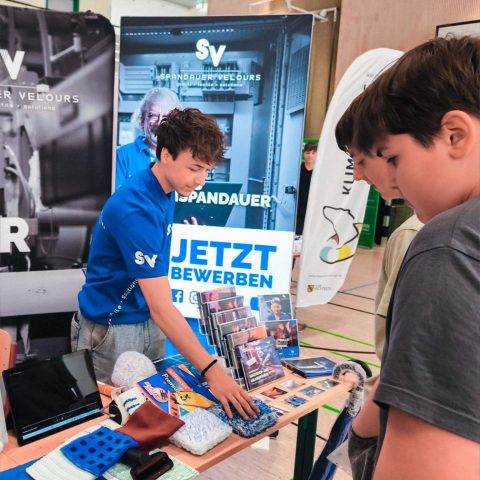 Der Auszubildende Vincent von Spandauer Velours erklärt interessierten Schülern die verschiedenen Textilprodukte am Messestand. Im Hintergrund ist ein Schild mit der Aufschrift „Jetzt bewerben“ zu sehen.