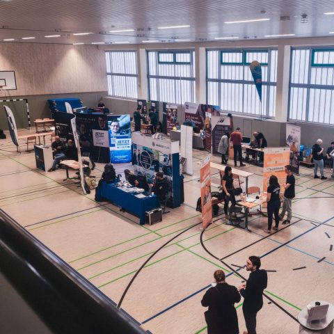 Vogelperspektive auf die gut besuchte Berufsorientierungsmesse in der Turnhalle der Oberschule Lugau. Man sieht verschiedene Messestände regionaler Unternehmen und zahlreiche Besucher im Austausch.