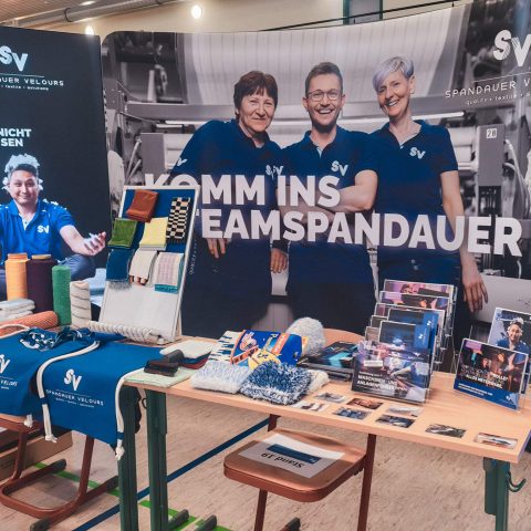 Nahaufnahme des Messestandes von Spandauer Velours mit Informationsflyern, Textilproben wie Velours-Stoffen und Reinigungstextilien sowie einem großen Hintergrundbanner mit lächelnden Teammitgliedern.
