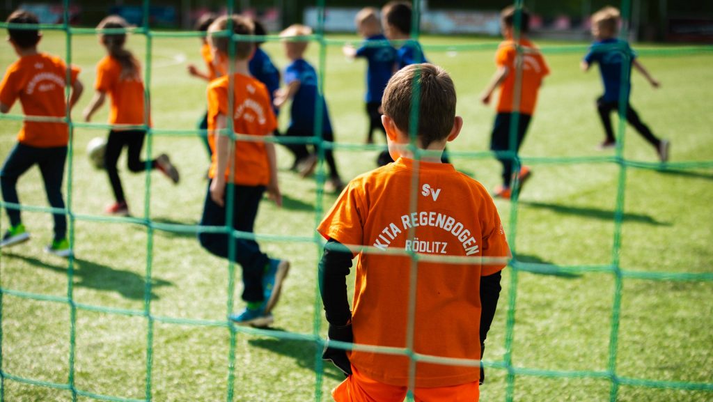 Kind von hinten fotografiert, er steht vor einem Fußballtor mit einem orangefarbenen Shirt mit dem Aufdruck "KITA REGENBOGEN", im Hintergrund weitere Kinder die Fußball spielen.