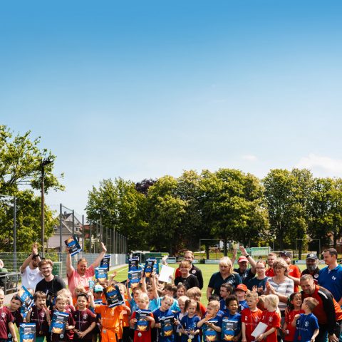 Großes Gruppenfoto aller teilnehmenden Kinder, Trainer und Sponsoren beim Kita-Cup 2026. Alle Beteiligten stehen jubelnd auf dem Fußballplatz unter blauem Himmel und halten stolz ihre Urkunden in die Kamera.