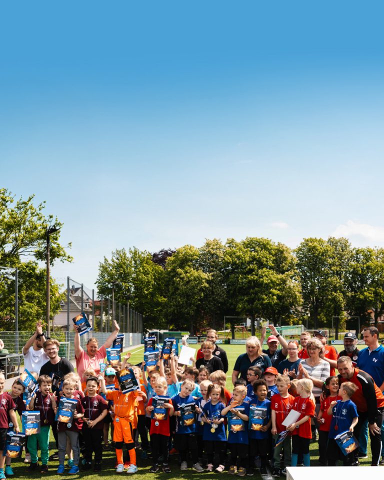 Großes Gruppenfoto aller teilnehmenden Kinder, Trainer und Sponsoren beim Kita-Cup 2026. Alle Beteiligten stehen jubelnd auf dem Fußballplatz unter blauem Himmel und halten stolz ihre Urkunden in die Kamera.