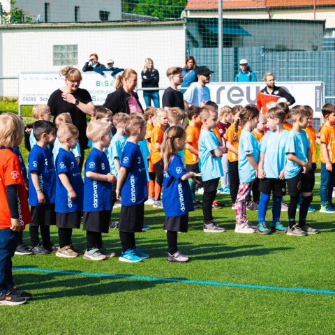 Aufstellung der Mannschaften beim Kita-Cup Kinderfußballturnier. Reihen von Kindern in blauen, hellblauen und orangen Trikots stehen nebeneinander auf dem grünen Rasen. Im Hintergrund stehen Trainer und Betreuer am Spielfeldrand bei sonnigem Wetter.