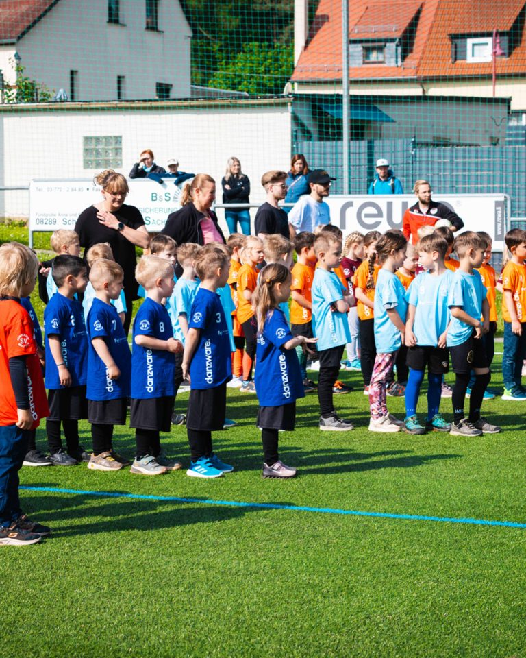 Aufstellung der Mannschaften beim Kita-Cup Kinderfußballturnier. Reihen von Kindern in blauen, hellblauen und orangen Trikots stehen nebeneinander auf dem grünen Rasen. Im Hintergrund stehen Trainer und Betreuer am Spielfeldrand bei sonnigem Wetter.