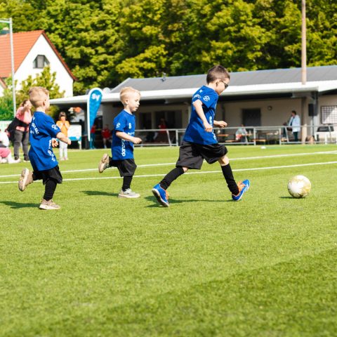 Dynamische Spielszene beim Kita-Cup. Drei Kinder in blauen Trikots des SV Heinrichsort/Rödlitz rennen auf dem Fußballplatz einem Fußball hinterher. Im Hintergrund sind Bäume und ein Vereinsheim zu sehen.