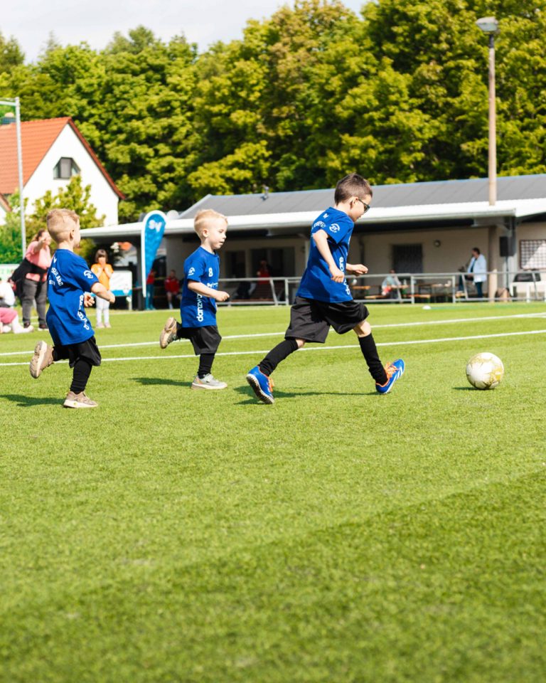 Dynamische Spielszene beim Kita-Cup. Drei Kinder in blauen Trikots des SV Heinrichsort/Rödlitz rennen auf dem Fußballplatz einem Fußball hinterher. Im Hintergrund sind Bäume und ein Vereinsheim zu sehen.