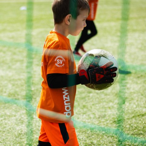 Alt-Text: Nahaufnahme eines jungen Torwarts beim Kita-Cup. Das Kind trägt ein oranges Trikot mit dem Logo des SV Heinrichsort/Rödlitz und Torwarthandschuhe. Es hält fokussiert einen Fußball im Arm und blickt durch das Tornetz auf das Spielfeld.