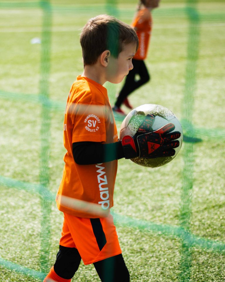Alt-Text: Nahaufnahme eines jungen Torwarts beim Kita-Cup. Das Kind trägt ein oranges Trikot mit dem Logo des SV Heinrichsort/Rödlitz und Torwarthandschuhe. Es hält fokussiert einen Fußball im Arm und blickt durch das Tornetz auf das Spielfeld.