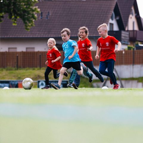 Spannender Zweikampf beim Kinderfußballturnier. Ein Kind in einem hellblauen Trikot dribbelt den Fußball, während drei Kinder in roten Trikots von Spandauer Velours versuchen, den Ball zu erobern. Wohnhäuser im Hintergrund.