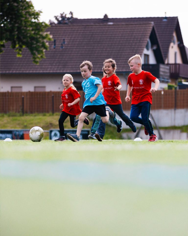 Spannender Zweikampf beim Kinderfußballturnier. Ein Kind in einem hellblauen Trikot dribbelt den Fußball, während drei Kinder in roten Trikots von Spandauer Velours versuchen, den Ball zu erobern. Wohnhäuser im Hintergrund.