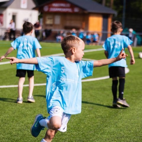 Ein glückliches Kind in einem hellblauen Trikot mit der Nummer 5 rennt mit ausgebreiteten Armen jubelnd über den Fußballplatz. Im Hintergrund laufen zwei Mitspieler.