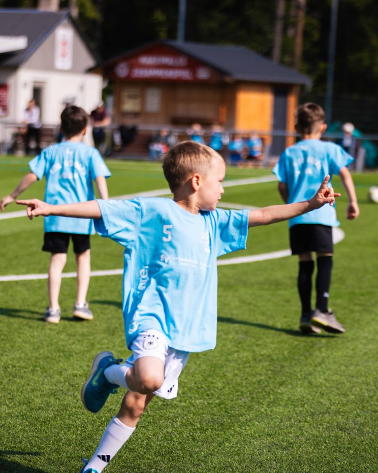 Ein glückliches Kind in einem hellblauen Trikot mit der Nummer 5 rennt mit ausgebreiteten Armen jubelnd über den Fußballplatz. Im Hintergrund laufen zwei Mitspieler.