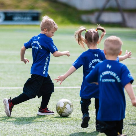 Drei Kinder der „Kita Knirpsenland Lichtenstein“ in dunkelblauen Trikots laufen gemeinsam über den Rasen. Ein Junge führt den Fußball eng am Fuß, die anderen laufen mit.