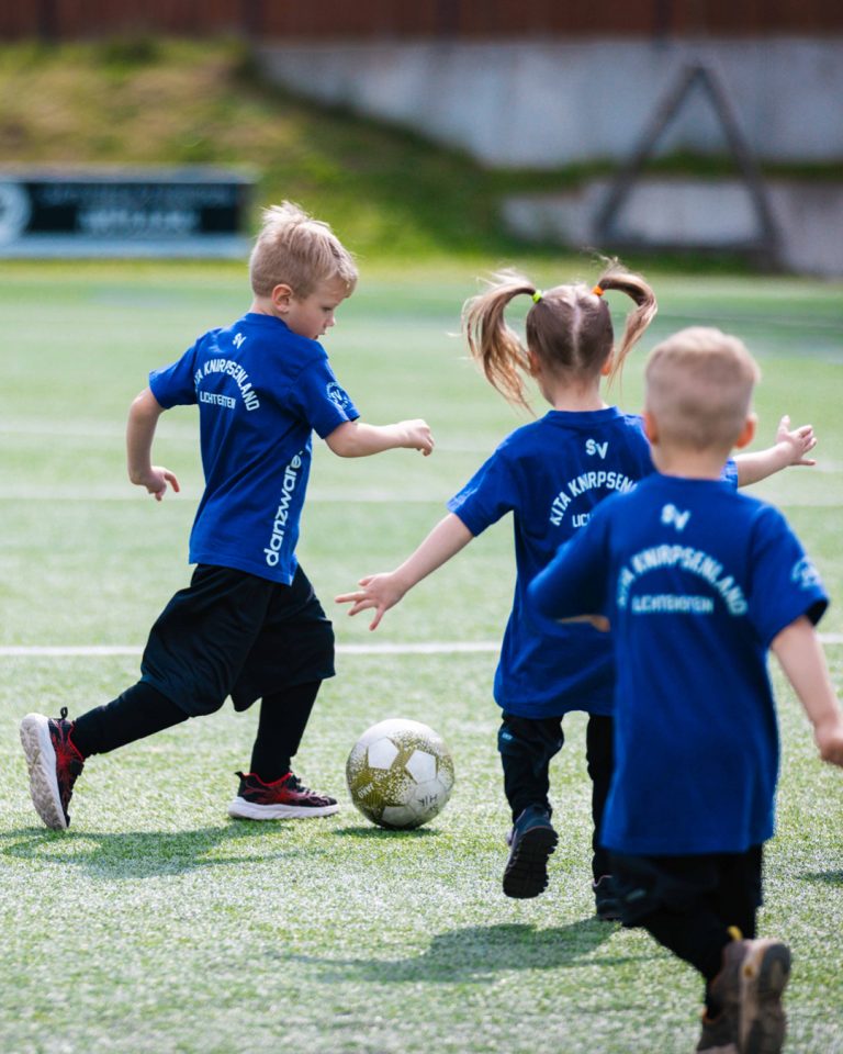Drei Kinder der „Kita Knirpsenland Lichtenstein“ in dunkelblauen Trikots laufen gemeinsam über den Rasen. Ein Junge führt den Fußball eng am Fuß, die anderen laufen mit.