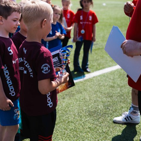 Ein kleiner Nachwuchsfußballer in einem weinroten Trikot der „Kita Schatzkiste“ hält stolz einen silbernen Pokal und eine Teilnehmerurkunde für den Kita-Cup in den Händen.