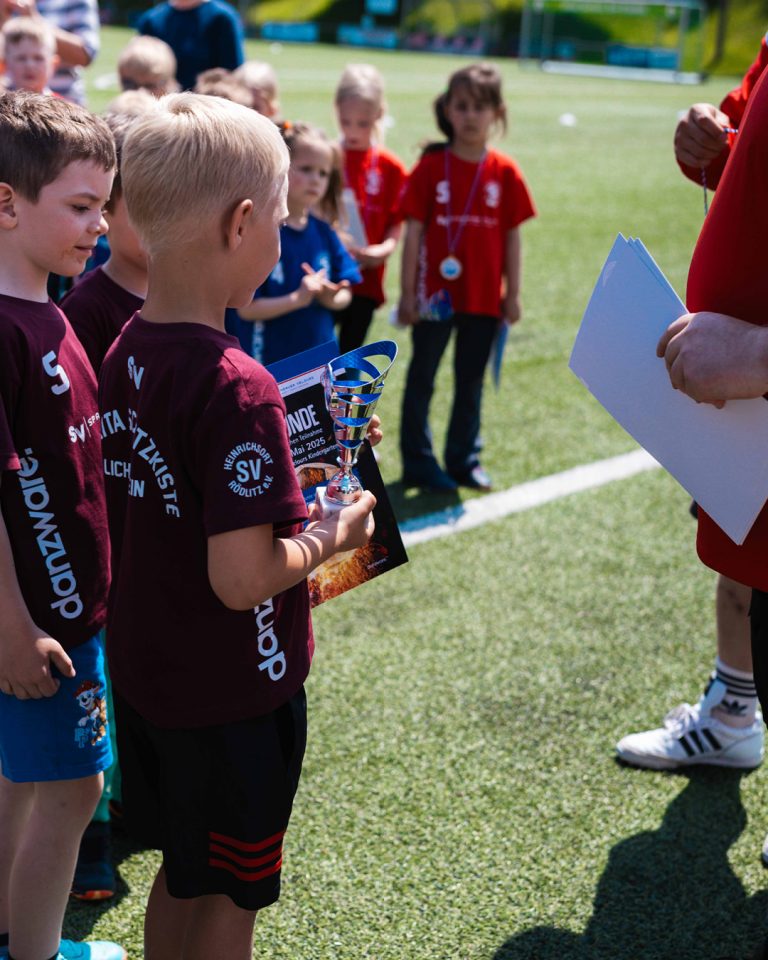 Ein kleiner Nachwuchsfußballer in einem weinroten Trikot der „Kita Schatzkiste“ hält stolz einen silbernen Pokal und eine Teilnehmerurkunde für den Kita-Cup in den Händen.