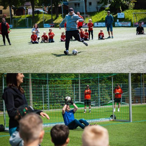 11. Ein erwachsener Trainer schießt während eines Kita-Fußballturniers auf dem Rasenplatz einen Ball, im Hintergrund sitzen applaudierende Kinder in roten Trikots und Zuschauer. Unten fängt ein sitzender Torwart den Ball vor einer Gruppe Kinder.
