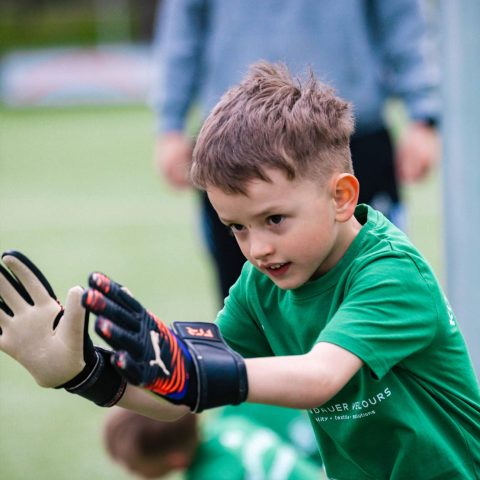 Ein konzentrierter Junge im grünen Trikot und mit Torwartandschuhen nimmt beim Kindergarten-Fußballturnier eine Abwehrhaltung auf dem Sportplatz ein.