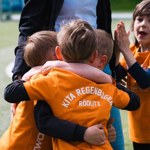 14. Drei glückliche Kinder im orangefarbenen Trikot der Kita Regenbogen Rödlitz umarmen sich jubelnd auf dem Fußballplatz.