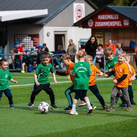 16. Eine Gruppe von Kindern in grünen und orangefarbenen Trikots kämpft eifrig um den Ball auf dem Fußballfeld der Kita Rappelkiste Hohndorf.