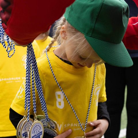 20. Ein stolzes Mädchen im gelben Trikot und grüner Kappe blickt lächelnd auf ihre gewonnene Goldmedaille beim Kita-Fußballturnier.