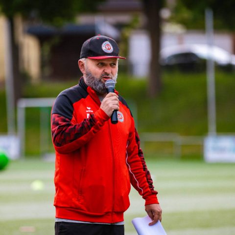 2. Ein Trainer oder Organisator des SV Heinrichsort/Rödlitz e.V. spricht mit einem Mikrofon und Notizen auf dem Fußballrasen zu den Teilnehmern.