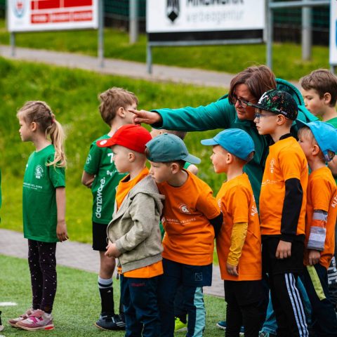 3. Eine Erzieherin in grüner Jacke gibt einer Gruppe von Kindergartenkindern in orangefarbenen T-Shirts Anweisungen am Spielfeldrand.
